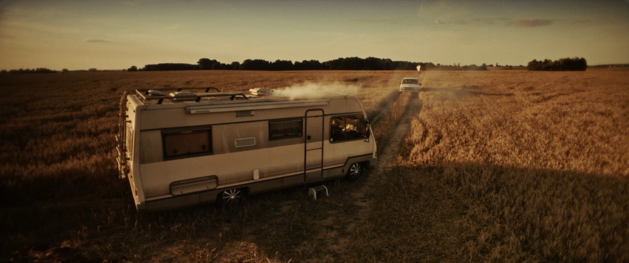 A vintage motorhome parked at the edge of a wheat field at golden hour, a car driving off in the distance - Amon Amarth